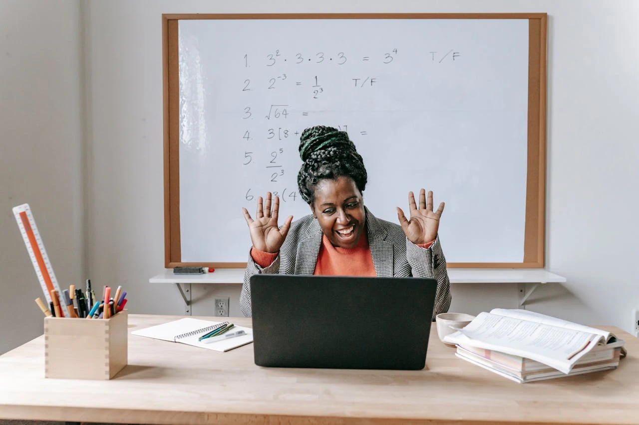 Professora sorridente em uma sala de aula com quadro branco, usando um notebook na mesa, enquanto ensina e interage com estudantes; ilustra como a educação online está transformando o acesso ao conhecimento no Brasil.